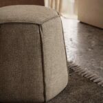 Een getextureerde, beige Angles Poef is geplaatst op een grijs franjekleed in een zacht verlichte kamer. De gespikkelde vloer en het doorschijnende gordijn op de achtergrond laten prachtig licht doorstromen.