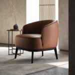 Een moderne woonkamer heeft een Megan Fauteuil met bruin leer en zwarte poten naast een ronde bijzettafel. Een minimalistische vaas met gedroogde takken staat op de tafel, terwijl zonlicht door een lattenwand filtert en artistieke schaduwen op de vloer creëert.