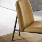 De Edison Armstoel is een moderne stoel met een mosterdgele, gewatteerde zitting en rugleuning, ondersteund door een zwart metalen frame. Hij staat elegant op een beige kleed met textuur tegen een lichtgrijze muur.
