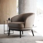 Een moderne beige Ruth Fauteuil met zwarte poten rust op een lichtgrijs kleed in een minimalistische kamer. Een klein zwart bijzettafeltje in de buurt bevat een kleine vaas met een groene plant. Het ontwerp van de kamer omvat houten muren, een grote betonnen pilaar en een uitnodigend raam.
