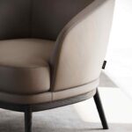 Een close-up van de Ruth Fauteuil, een moderne beige fauteuil met gebogen armleuningen en houten poten, staat elegant op een licht tapijt in een zonnige kamer. Een klein zwart label is netjes bevestigd aan de zijnaad.