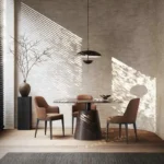 Een moderne eethoek met een ronde houten tafel van Anna Stoel - Houten Poten, drie bruine stoelen en een hanglamp. Een hoge vaas met takken staat op een voetstuk tegen een muur met structuur. Zonlicht stroomt door jaloezieën en creëert ingewikkelde patronen op muren en vloer.