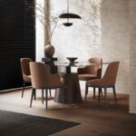 Een moderne eethoek heeft een ronde glazen tafel met vier bruine stoelen, waaronder een Anna Stoel Met Armleuning - Houten Poten. Een grote vaas met takken vangt natuurlijk licht via een raam met jaloezieën. Boven het hoofd accentueert een zwarte hanglamp de grijze muur met textuur.