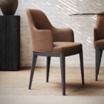 De Anna Stoel Met Armleuning - Houten Poten toont een modern design met een lichtbruine gestoffeerde zitting en rugleuning, ondersteund door stevige houten poten. Het staat elegant op een lichte houten vloer, aangevuld met de getextureerde bakstenen muur en hints van een glazen tafel en vaas op de achtergrond.