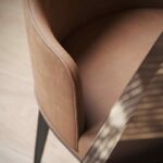 Close-up van een zachtbruine gestoffeerde stoel met een gebogen rug en zwarte houten poten, geplaatst bij een houten oppervlak. Deze Anna Stoel Met Armleuning - Houten Poten toont op prachtige wijze de elegante textuur en het minimalistische ontwerp van de stoel, waarbij comfort en stijl worden gecombineerd.