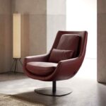 Een eigentijdse ELBA Fauteuil, gemaakt van rood leer, compleet met een bijpassend kussen, rust elegant op een betonnen vloer. Een hoge staande lamp werpt een zachte gloed tegen de neutrale beige en witte muren.