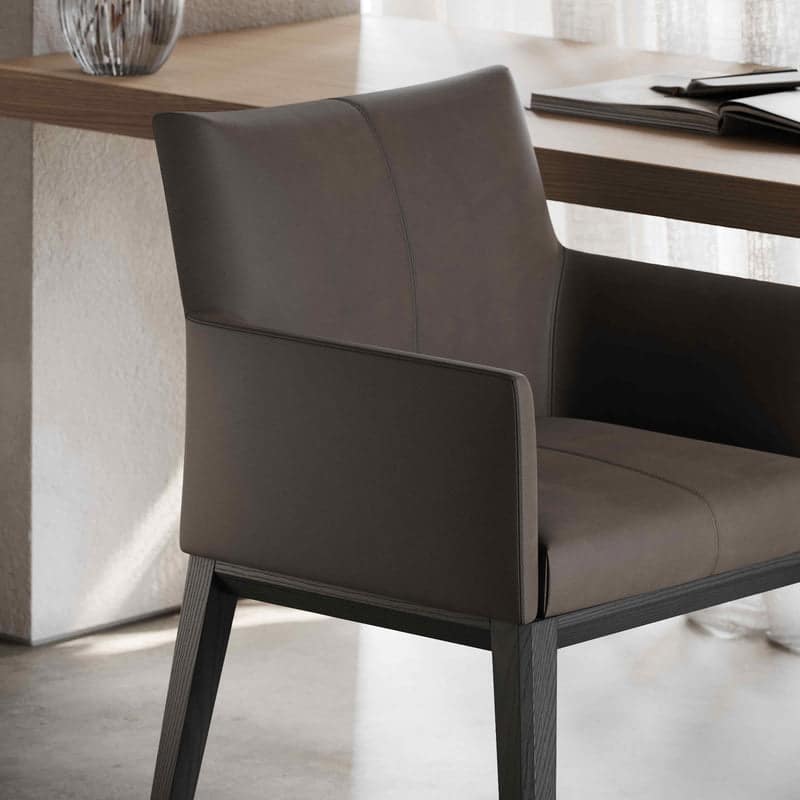 De Carter Stoel verbetert elk modern kantoor met zijn elegante design, met strak donkerbruin leer en houten poten. Geplaatst naast een houten bureau met een notitieboek en pen, bevordert het de productiviteit terwijl zacht natuurlijk licht door de gordijnen op de achtergrond stroomt.