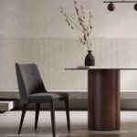De elegante eethoek benadrukt de moderne Louise Stoel stoel in donkergrijs, geplaatst rond een ronde tafel met een bruine cilindrische basis. De ambiance wordt versterkt door minimalistische verlichting en een getextureerde muur, terwijl droge takken in een vaas een organische toets aan de setting geven.