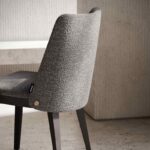 De Louise Stoel, een moderne stoel met een strak houten frame en grijze bekleding met textuur, staat tegen betonnen muren en vloeren. Het heeft een minimalistisch design met een zwart-gouden label op de rugleuning.