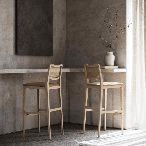 De minimalistische ruimte omvat een hoge tafel gecombineerd met twee Eva Barkruk houten barkrukken, terwijl een witte vaas met takken elegantie toevoegt. Natuurlijk licht stroomt door transparante gordijnen en werpt zachte schaduwen op de grijze getextureerde muren en vloer.