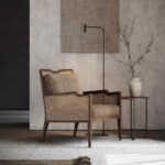 Een Eva Fauteuil, met een geweven rugleuning en een gewatteerde zitting, is gezellig neergezet bij een minimalistische bijzettafel. Een slanke staande lamp complementeert een vaas gevuld met takjes, tegen een getextureerde muur met een abstract canvas erboven.