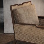 Close-up van de Eva Fauteuil, met een mid-century modern houten frame en geweven rieten rugleuning. Een beige kussen met textuur rust uitnodigend tegen de rugleuning, wat de neutrale muur en vloer complementeert.