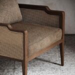 De Eva Fauteuil is een houten fauteuil met een rugleuning en armleuningen van rotan en een zitting met beige kussens. De fauteuil staat op een gespikkelde grijze vloer tegen een muur met structuur, waardoor de warme, rustieke sfeer wordt versterkt.