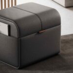 De Adley Poef S is een zwarte leren ottoman met een strak design en zichtbare stiksels. Het bevat een verborgen opbergruimte met een bruine leren tab, waar een tijdschrift elegant uit piept terwijl het op een donker tapijt rust.