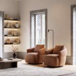 De gezellige, moderne woonkamer toont een paar bruine fauteuils van Alice Fauteuil, aangevuld met een strakke staande lamp en een licht tapijt. Zonlicht stroomt door grote ramen en verlicht een aan de muur bevestigde plank met decoratie. Een houten salontafel toont een vaas met gedroogde bloemen.