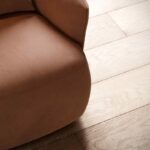 Een close-up van de Alice Fauteuil, met zachte, ronde armleuningen van bruin leer, staat op een lichte houten vloer met zichtbare nerf. Hierdoor ontstaat een gezellige, minimalistische esthetiek met de nadruk op textuur en materiaal.