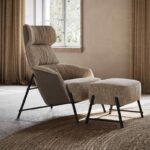 Een gezellige, getextureerde fauteuil met bijpassende voetenbank van Hubert Voetensteun rust op een rond geweven tapijt. De stoel staat op strakke zwarte metalen poten, terwijl zonlicht door beige-gordijnen ramen stroomt en een zachte gloed over de neutrale kamer werpt.