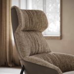 Een close-up van de getextureerde Hubert Fauteuil in beige toont het comfortabele ontwerp met hoge rugleuning, geplaatst bij een raam met een neutraal gordijn voor een gezellige sfeer.