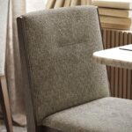 Een close-up van de Frank Stoel eetkamerstoel toont de grijze bekleding met textuur en het verfijnde ontwerp. De stoel heeft een hoge rugleuning en donkere houten poten, met gestapelde boeken en een tafel met marmeren blad die de elegante scène op de achtergrond completeren.