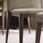 De afbeelding toont een houten stoel van Frank Stoel met een getextureerde groenbeige gestoffeerde zitting en donkere houten poten. Een andere soortgelijke stoel is gedeeltelijk zichtbaar ernaast op de lichtgrijze betegelde vloer, wat de esthetiek van het ontwerp weerspiegelt.