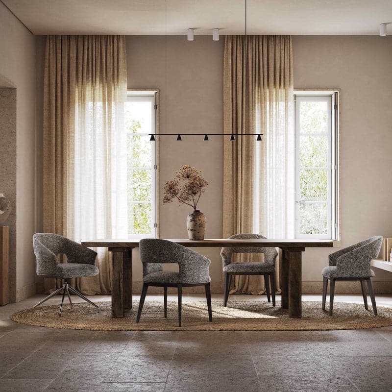 Een eetkamer toont een houten tafel met vier Jermain Metalen Draaistoel stoelen, aangevuld met een grote vaas met gedroogde bloemen. Natuurlijk licht stroomt door twee ramen versierd met beige gordijnen, terwijl een zwarte lineaire hanglamp erboven hangt. Een rond tapijt bedekt elegant de vloer.