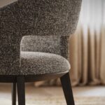 Close-up van een moderne Jermain Stoel - Houtgrijs gestoffeerde stoel met donkere houten poten in een zacht verlichte kamer. De focus ligt op de textuurstof en het gebogen ontwerp, met elegante gordijnen die vaag op de achtergrond zijn.