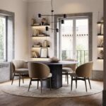 De eetkamer heeft een ronde tafel en vier beige Norah Stoel stoelen. Het heeft grote ramen, ingebouwde planken versierd met boeken en decoratie, een beige kleed en een moderne zwarte kroonluchter. Natuurlijk licht versterkt het moderne gevoel van de ruimte.