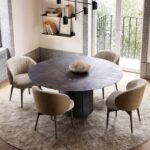 Een moderne eetkamer toont een donkere ronde Norah Stoel-tafel op een licht tapijt, omringd door vier gestoffeerde stoelen en bekroond met een decoratieve schaal. Boekenplanken sieren de muur, terwijl een groot raam natuurlijk licht verwelkomt.