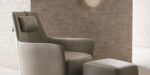 Een Alexander Fauteuil in lichtgrijs, met bijpassende ottoman, staat tegen een getextureerde bakstenen muur. Een ronde wandlamp erboven zorgt voor een zachte gloed, wat een gezellige en minimalistische ambiance creëert.