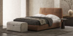In een moderne slaapkamer is een bruin gestoffeerd bed versierd met een donkergrijze deken en witte kussens. Een Adley Poef L voegt elegantie toe aan het voeteneind van het bed, met een minimalistisch bijzettafeltje met een vaas met droge takken tegen een lichte bakstenen muur.