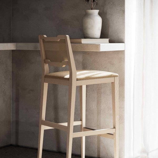 Een houten kinderstoel van Eva Barkruk met een gevlochten rugleuning staat naast een witte tafel aan de muur, waar een witte vaas met gedroogde bloemen en een boek op staan. Zonlicht stroomt door het gordijn en werpt zachte schaduwen.