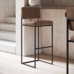 De Gram Barkruk is een moderne barkruk met een bruine, gewatteerde zitting en rugleuning, op een strak zwart metalen frame. Hij combineert elegant met de lichte houten vloer en verbetert de uitstraling van de nabijgelegen neutrale trappen.