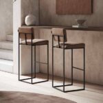Een modern interieur met twee Gram Barkruk krukken met bruine, gestoffeerde zittingen en zwarte metalen frames aan een minimalistische bar, versierd met een vaas en kom in neutrale tinten. Zachte verlichting voegt warmte toe aan de setting.