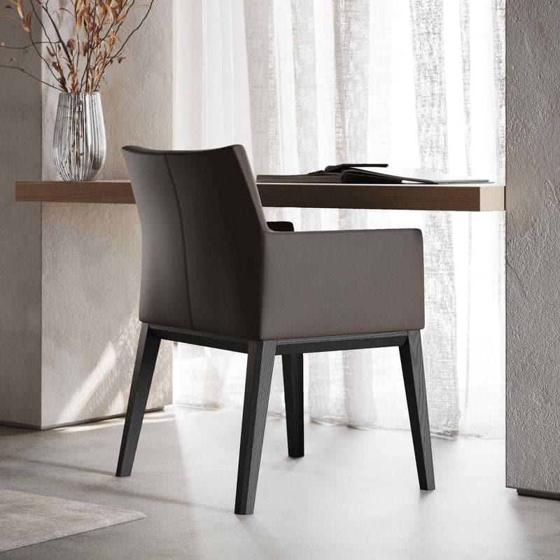 Een moderne, donkergrijze Carter Stoel stoel staat naast een houten bureau voor een raam met doorschijnende witte gordijnen die zacht licht filteren. Het bureau is versierd met een vaas met gedroogde takken.