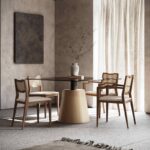 Een moderne eetkamer toont een ronde glazen tafel met vier Eva Stoel-stoelen. Een vaas vol takken siert de tafel, terwijl natuurlijk licht zachtjes door de transparante gordijnen filtert, wat de warme, minimalistische ambiance in neutrale tinten benadrukt.