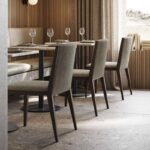 Een moderne eetruimte toont ontwerpen van Frank Stoel, met houten stoelen en tafels voor vier. Elegant servies en wijnglazen sieren de tafels. Natuurlijk licht filtert door de ramen en verlicht het stijlvol ingerichte interieur.