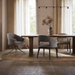 Een eethoek heeft een Jermain Stoel - Hout houten tafel met moderne grijze stoelen op een geweven tapijt. Grote ramen met doorschijnende beige gordijnen laten natuurlijk licht binnen en de tafel is versierd met een vaas met gedroogde bloemen. Een minimalistische hanglamp hangt er elegant boven.