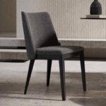 De moderne donkergrijze Louise Stoel, elegant gestoffeerd, staat op zwarte houten poten op een betonnen vloer. Een getextureerde muur en een plank met een keramische vaas dragen bij aan de minimalistische elegantie van de scène.