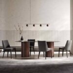 Een moderne eetkamer is geheel in de stijl van Louise Stoel ingericht met een ronde glazen tafel op een tapijt met patroon, vier grijze stoelen, een vaas met takken, een minimalistische hanglamp met vijf bruine bollen en muren met een beige structuur.
