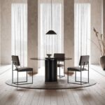 Een moderne eetkamer toont drie Gram Stoel-stoelen rond een donkere ronde tafel, verlicht door een hanglamp erboven. De ruimte heeft hoge ramen met doorzichtige gordijnen en een minimalistisch decor, versterkt door natuurlijk licht dat op een kleine kom op de tafel valt.