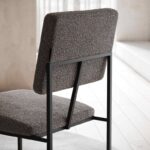 De Gram Stoel stoel heeft een zitting en rugleuning van getextureerde grijze stof en een strak zwart metalen frame. Het geheel staat elegant tegen een zacht verlichte, neutrale achtergrond.