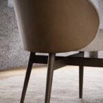 Een close-up van de Norah Stoel, een moderne stoel met bekleding van bruin leer en beige stof, op strakke houten poten op een crèmekleurig tapijt tegen een grijze muur.