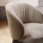 De Norah Stoel, een moderne fauteuil met een gebogen rugleuning, staat op een hardhouten vloer met een structuurstof in neutrale tinten en een mix van gladde en patroonstoffen voor een elegante, eigentijdse look.