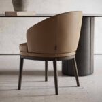 De Norah Stoel, met zijn moderne beige bekleding, gebogen rugleuning en donkere houten poten, vormt een mooie aanvulling op de minimalistische, zwarte, cilindervormige tafel ernaast. Hierop staan een boek en een vaas, tegen een neutrale muur, waardoor een elegante en serene sfeer ontstaat.