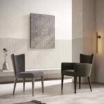 Een minimalistische kamer toont twee moderne gestoffeerde stoelen, een in grijs en de andere in groen, op een lichte betonnen vloer. Een abstract schilderij met textuur siert een tweekleurige muur, terwijl de Vianna Stoel wandlamp warme verlichting biedt, wat de serene, hedendaagse sfeer versterkt.