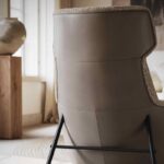 De Hubert Fauteuil is een stijlvolle stoel met hoge rugleuning, beige leren en getextureerde stoffen accenten, op een strak zwart metalen frame. Een houten tafel en een grote vaas bij zonovergoten gordijnen voegen warmte toe aan de kamer.