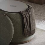 Een groene Avery Poef met een ronde, platte bovenkant en een gleuf in het midden toont een bruine franje eroverheen gedrapeerd. Een bijpassend kussen of deksel staat ernaast op een getextureerde vloer, met een bruin houten oppervlak subtiel op de achtergrond.