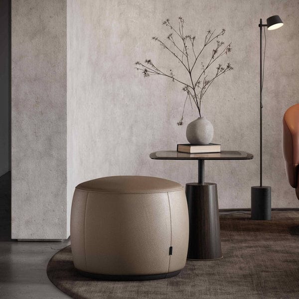 Een minimalistische kamer omvat een ronde beige Rachel S Poef, een kleine tafel met een vaas met gedroogde takken, een staande vloerlamp en een bruine fauteuil. De getextureerde muur draagt bij aan het gezellige moderne gevoel.