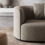 Een moderne Chloe 2-zitsbank met een gebogen design in beige structuurstof staat elegant in een minimalistische kamer. Het is versierd met een bijpassend kussen, aangevuld met een neutraal getint tapijt eronder en vergezeld door een artistiek steenachtig aardewerkstuk op de achtergrond.