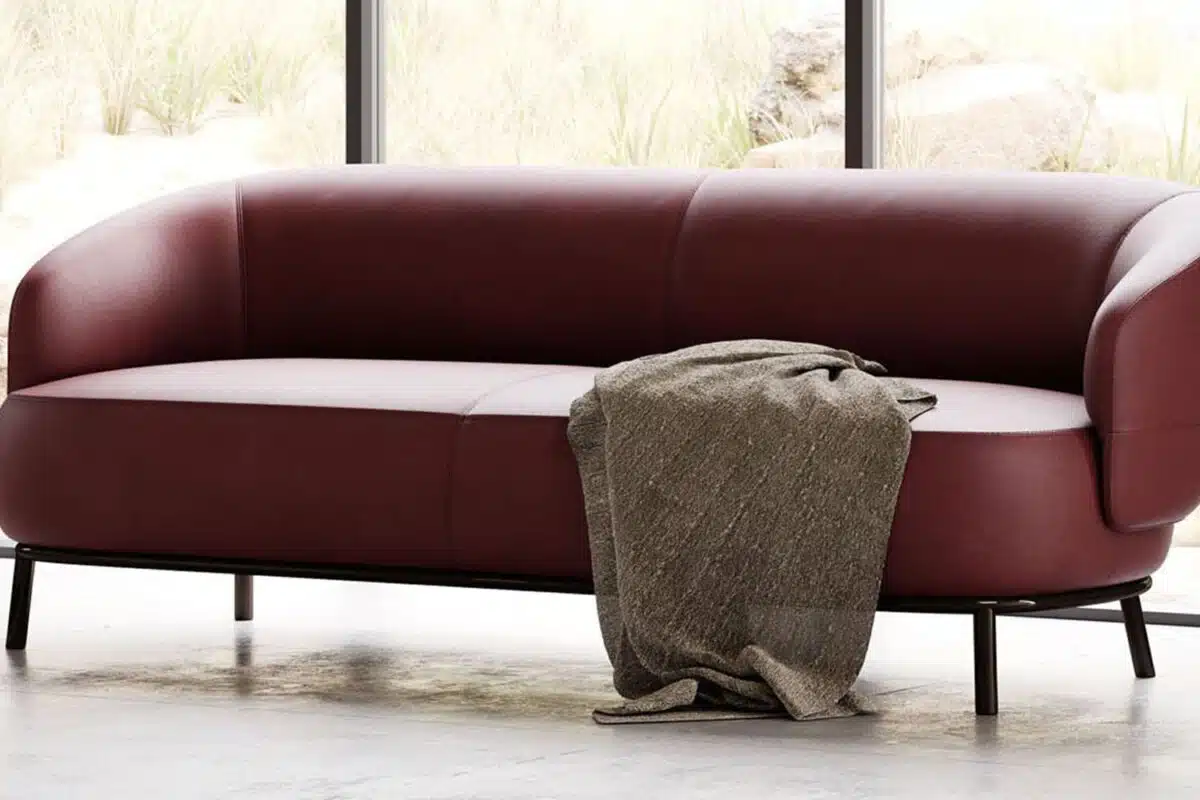 De kastanjebruine Juliet 2-zitsbank met zwarte poten staat elegant in een lichte kamer met grote ramen. Een grijze getextureerde deken is over de arm gedrapeerd, terwijl de achtergrond een rustig uitzicht op rotsen en planten buiten laat zien.